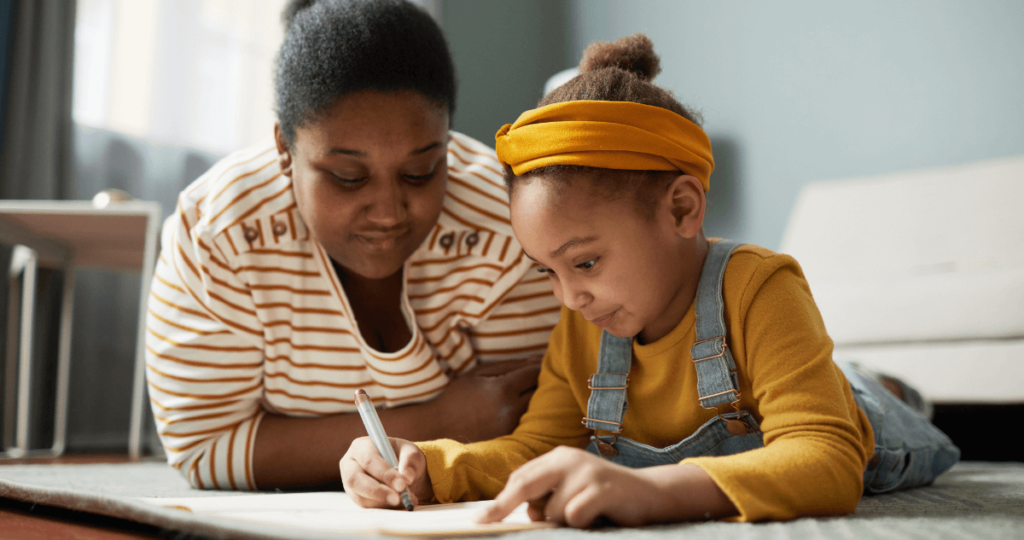 Mother and daughter laying on the ground as the mother watches the daughter draw or write on a piece of paper.