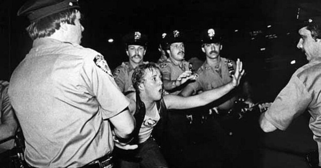 A black and white image from the Stonewall riots of a young person surrounded by police officers who are reaching for them.