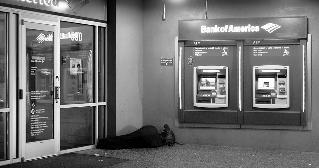 Black and white image of a homeless person asleep in a sleeping bag next to a Bank of America.