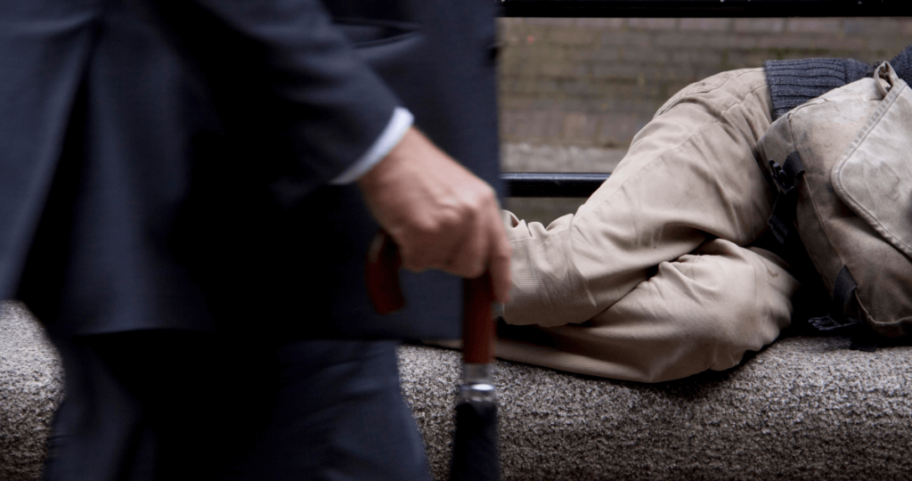 Businessman on a suit walking past a man sleeping on a bench.