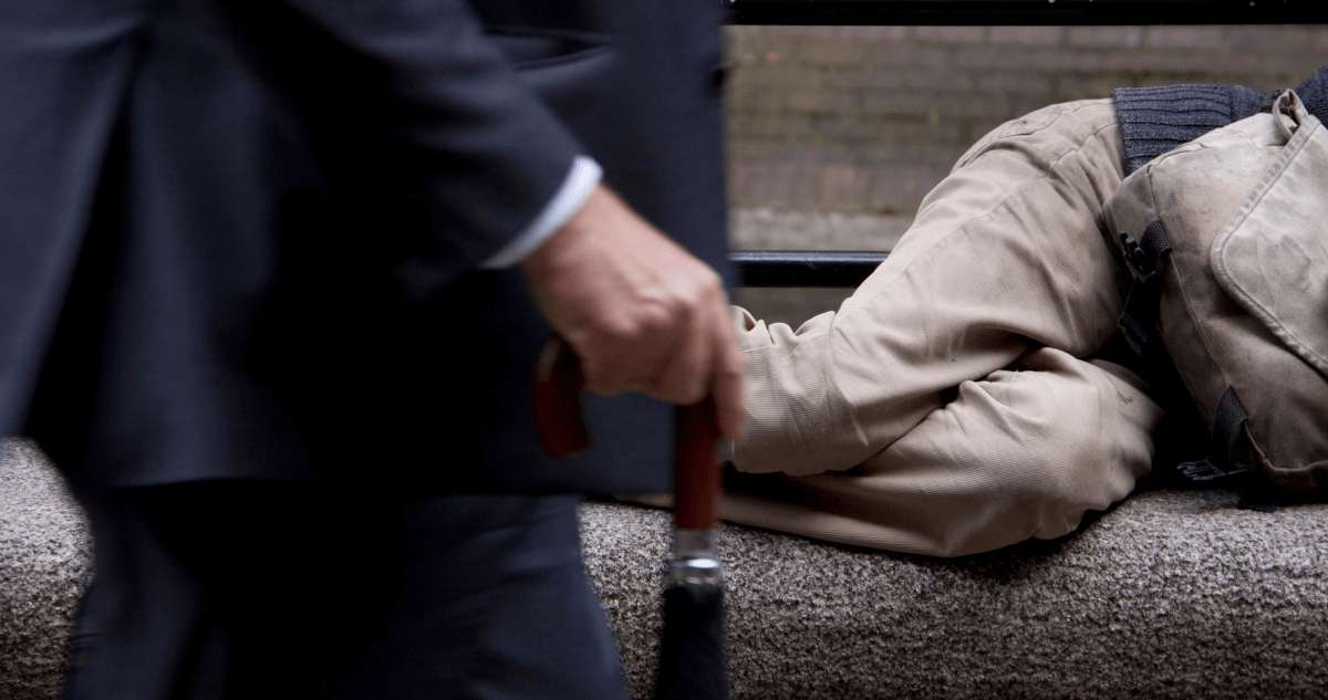 Businessman on a suit walking past a man sleeping on a bench.