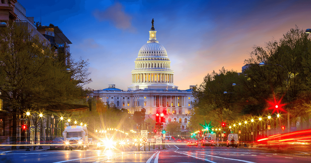 View of the US Capitol from a busy street at dusk, with headlights and traffic lights glowing in the foreground.