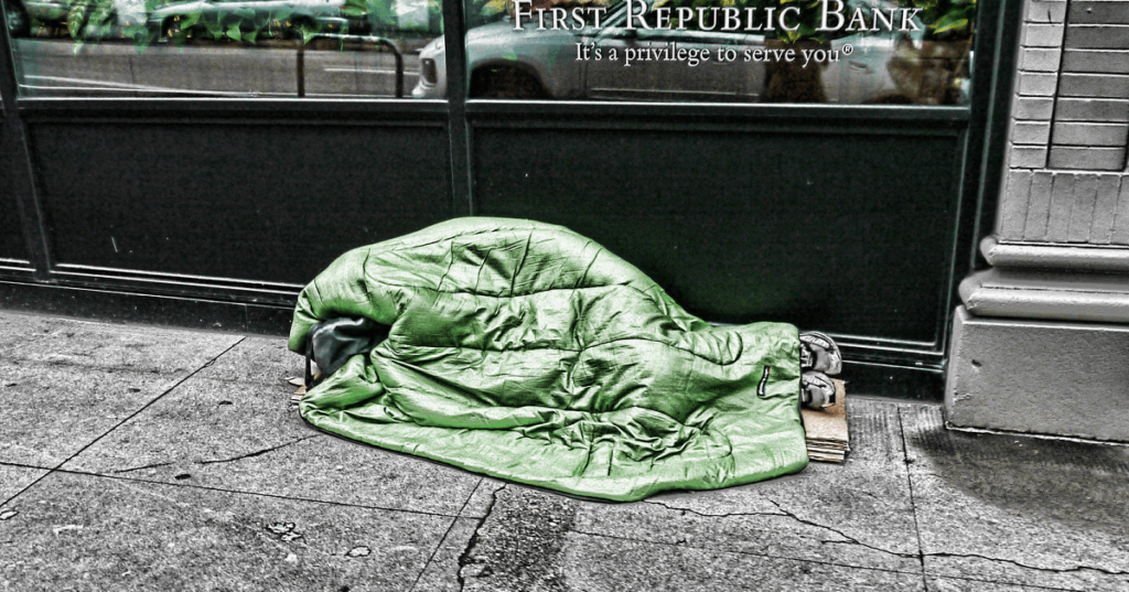 Image of a homeless person laying under a sleeping bag on a sidewalk.