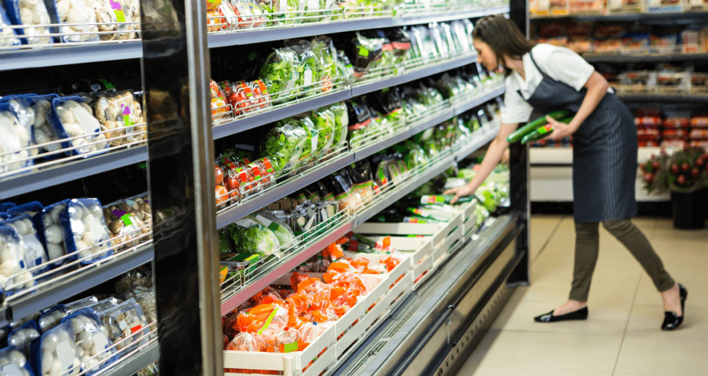 Woman wearing an apron at a supermarket produce section restocking shelves.